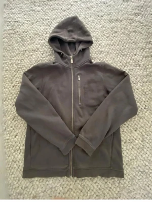 Mörkgrå zip hoodie från Sail Racing - Mörkgrå hoodie från Sail Racing med dragkedja hela vägen, huva och bröstficka med zip. Tillverkad i mjukt material, perfekt för chill dagar. Diskret logga på bröstet och två sidofickor. Snygg och enkel design som funkar till det mesta.
