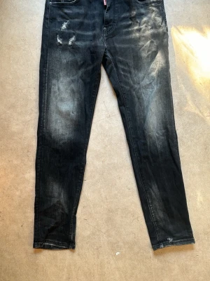 DSQ jeans - Säljer ett par svarta jeans med slitningar och tvättade detaljer för en cool, avslappnad look. Jeansen har raka ben, fem fickor och synliga slitningar både fram och bak. Perfekta för dig som gillar streetwear och en trendig stil.