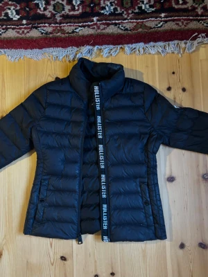 Hollister Ultimate Down Jacket XS – svart slim fit puffer / höstjacka - Säljer en svart Hollister-jacka från Hollister’s Ultimate Down Collection i storlek XS.  Jackan har en väldigt snygg och figurnära slim fit-passform som sitter smickrande utan att kännas klumpig. Den är lätt, bekväm och perfekt som höstjacka, vår jacka eller tunnare vinterjacka beroende på väder. Modellen har blank svart puffer-design, hög krage, Hollister-logga längs dragkedjan och logopatch på ärmen.  Passar perfekt om man vill ha en enkel men snygg vardagsjacka som ändå känns lite mer sportig