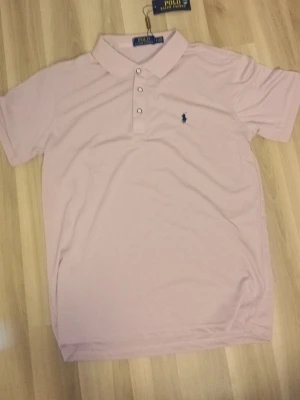 Ljusrosa polotopp från Polo Ralph Lauren - Säljer en ljusrosa polotopp från Polo Ralph Lauren med klassisk krage och tre knappar framtill. Toppen har korta ärmar och är tillverkad i mjuk bomull. Den ikoniska loggan är broderad på bröstet.