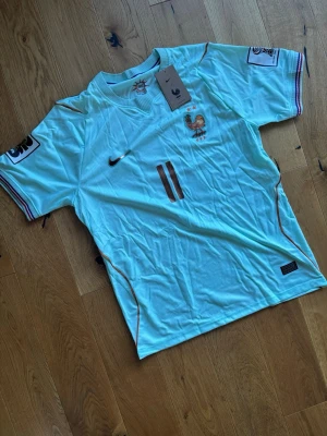 Frankrike fotbollströja Nike Olise - Ljusblå fotbollströja från Nike med Frankrikes landslagsemblem och nummer 11 på bröstet. Namnet Olise och nummer 11 i bronsfärg på ryggen. Korta ärmar med randiga detaljer i blått, vitt och rött. Tillverkad i lätt och ventilerande material, perfekt för match eller träning. Helt oanvänd 