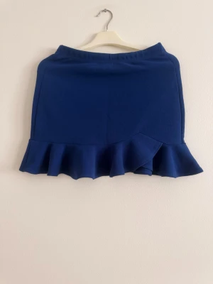 Blå volangkjol med struktur - Snygg blå kortkjol med volangkant nedtill. Kjolen har en enkel, stilren design och är tillverkad i ett strukturerat material som ger en modern känsla. Perfekt att matcha med en enkel topp för en trendig look.