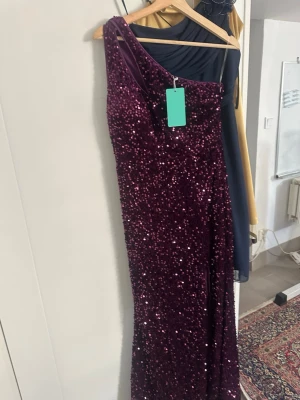 Vinröd glittrig oneshoulderklänning - Säljer en vinröd långklänning med glittriga paljetter och oneshoulder-design. Klänningen har en elegant och figurnära passform, perfekt för speciella tillfällen. Materialet är troligtvis polyester och den gnistrar snyggt i ljuset.