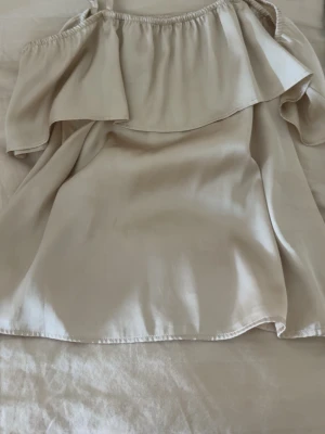 Beige offshoulder topp från Lindex - Säljer en beige offshoulder topp från Lindex med volangdetalj över bysten och tunna axelband. Toppen har en luftig passform och är perfekt för varma dagar. Materialet känns mjukt och lätt mot huden.