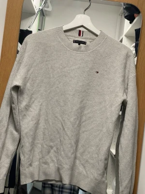 Ljusgrå stickad tröja Tommy Hilfiger - Snygg ljusgrå stickad tröja från Tommy Hilfiger med diskret logga broderad. Tröjan har rund halsringning, ribbade muddar och är tillverkad i ett mjukt, tunt stickat material. Perfekt för lager på lager och enkel att matcha. Passar S/M
