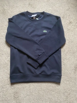 Mörkblå sweatshirt från Lacoste - Klassisk mörkblå sweatshirt från Lacoste med rund halsringning och det ikoniska gröna krokodilmärket broderat på bröstet. Tröjan har ribbade muddar vid ärmslut och nederkant. Perfekt för en clean och stilren look.
