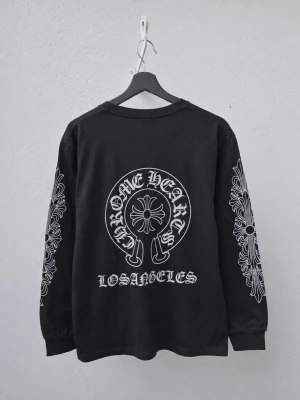 Svart långärmad topp Chrome Hearts - Svart långärmad topp från Chrome Hearts med stora vita tryck på ryggen och båda ärmarna. Motivet inkluderar texten 'Chrome Hearts Los Angeles' och grafiska detaljer längs ärmarna. Tillverkad i bomull med relaxed fit och rund hals.