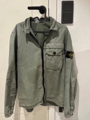 Grön overshirt från Stone Island - Snygg grön overshirt från Stone Island med klassisk patch på ärmen. Overshirten har en bröstficka med knapp, dragkedja framtill och avslappnad passform. Tillverkad i bomull och perfekt för lager på lager-stil.
