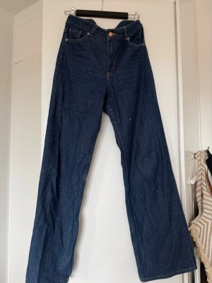 Mörkblåa vida jeans med hög midja - Säljer ett par mörkblå jeans med hög midja och vida ben. Klassisk femficksmodell med knapp och dragkedja framtill. Jeansen har en avslappnad passform och är tillverkade i denim. Perfekta för en trendig och bekväm stil.