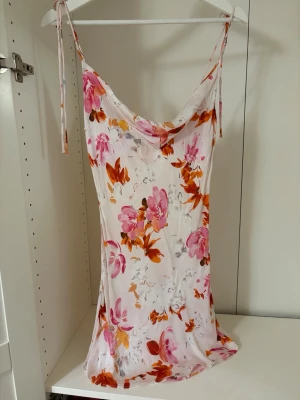Blommig sommarklänning från Zara - Söt kort klänning från Zara med blommigt mönster i rosa, orange och rött på vitt. Klänningen har tunna axelband som knyts och en lätt draperad urringning. 