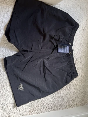 Svarta shorts från Prada med snörning - Säljer ett par svarta shorts från Prada med elastisk midja och snörning. Shortsen har en klassisk passform och en diskret Prada-logga på vänster ben. Perfekta för sommaren och tillverkade i ett lätt syntetmaterial.