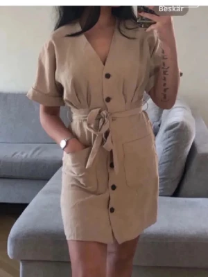 Zara sommarklänning❤️ - Stilren beige kortklänning med v-ringning, knappar framtill och knytband i midjan. Klänningen har korta ärmar med uppvik och stora fickor framtill. 