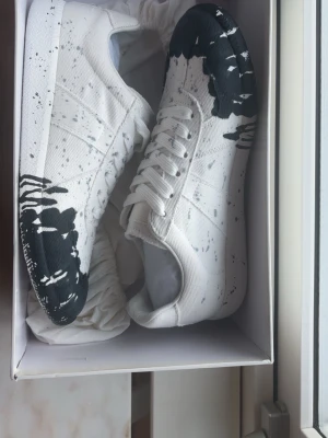 Maison Margiela vita sneakers med mörkblå färgstänk - Unika vita sneakers från Maison Margiela med svart färgstänksdesign på tå och sula. Skorna har klassisk låg profil, vita snören och är tillverkade i canvas med gummisula. Perfekt för dig som gillar statement-plagg och vill sticka ut med din stil.