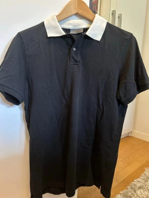 Svart polotopp med vit krage från Karl Lagerfeld - Säljer en svart polotopp från Karl Lagerfeld med klassisk vit krage och knappar framtill. Toppen är kortärmad och har en normal passform, perfekt för dig som gillar stilrena och enkla plagg. Materialet är mjuk bomull som känns skönt mot huden.