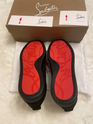 Christian Louboutin svarta sneakers - Svarta sneakers från Christian Louboutin med klassisk röd sula och svarta nitar på tån. Skorna har snörning, låg profil och är tillverkade i mocka med detaljer i läder. Perfekta för dig som vill sticka ut med en edgy och lyxig stil.