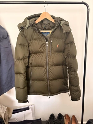 Olivgrön pufferjacka från Polo Ralph Lauren - Snygg olivgrön pufferjacka från Polo Ralph Lauren med avtagbar huva, dragkedja framtill och flera fickor. Storlek S passar längden 175-185