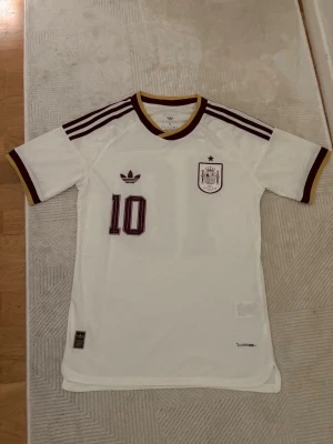 Vit Spanien fotbollströja Adidas - Snygg vit Spanien fotbollströja från Adidas med vinröda detaljer och nummer 10. Tröjan har Lamine Yamal tryckt på ryggen, klubbmärke på bröstet och klassiska Adidas-ränder på axlarna. Perfekt för dig som älskar fotboll och vill sticka ut på planen.