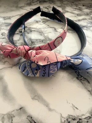 Söta hårdiadem med rosett, 2-pack - Två hårdiadem i plast klädda med tyg, ett i rosa och ett i ljusblått, båda med lekfullt mönster och knutna rosetter framtill. Perfekta för att ge en färgglad och gullig touch till din frisyr.