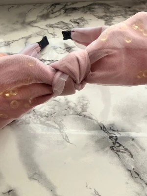 Rosa hårband med knut och guldprickar - Supersött hårband i ljusrosa tyg med en stor knut framtill och små guldiga prickar som detalj. Materialet är tunt och lätt, troligen polyester eller liknande. Perfekt accessoar för att piffa upp frisyren med en trendig touch.