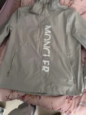Grå vindjacka från Moncler - Snygg grå vindjacka från Moncler med stor vit logga längs dragkedjan och klassisk Moncler-patch på ärmen. Jackan har huva, två sidofickor med dragkedja och är tillverkad i ett lätt, vattenavvisande material. Perfekt för blåsiga dagar.jackan är även helt ny aldrig använt 