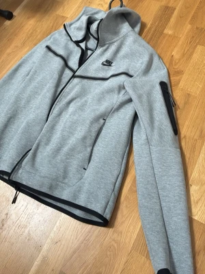 Nike tech fleece  - Snygg grå hoodie från Nike med svarta dragkedjor och detaljer. Har huva, två sidofickor och en extra ficka med dragkedja på ärmen. Perfekt för en sportig och avslappnad stil. Materialet är mjukt och stretchigt för extra komfort.