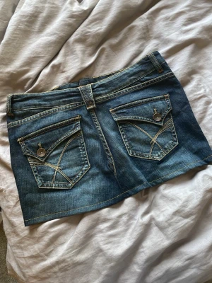 Blå jeanskjol med fickor - Säljer en blå jeanskjol i kort modell med två stora bakfickor och slitna detaljer framtill. Kjolen har klassisk jeanslook med bälteshällor och kontrastsömmar. Perfekt för dig som gillar denim och vill ha en cool, avslappnad stil.