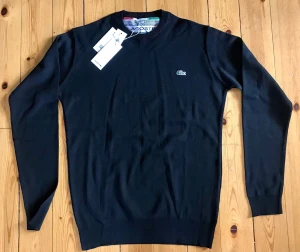 Svart tröja från Lacoste - Klassisk svart tröja från Lacoste med rund halsringning och diskret krokodillogga på bröstet. Långärmad modell i mjukt material, perfekt för en stilren look. Ribbstickade muddar vid ärmslut och nederkant.