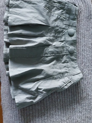Grå skort med knappar och fickor - Snygg grå skort med två stora knappar framtill och flera fickor både fram och bak. Plisserad design som ger en kjol-look men med shortskomfort. Tillverkad i ett mjukt bomullstyg, perfekt för en avslappnad och trendig stil.