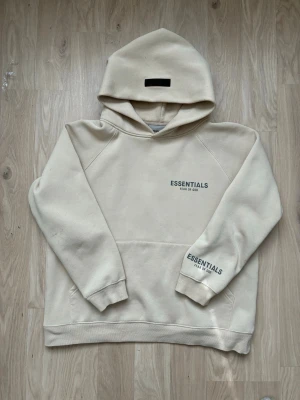 Beige Essentials Fear of God hoodie - Snygg beige hoodie från Essentials Fear of God med svart logga på bröstet och ärmen. Hoodien har en stor huva, känguruficka och ribbade muddar. Tillverkad i mjukt bomullsmaterial som känns riktigt skönt mot huden. Perfekt för en avslappnad och trendig look.