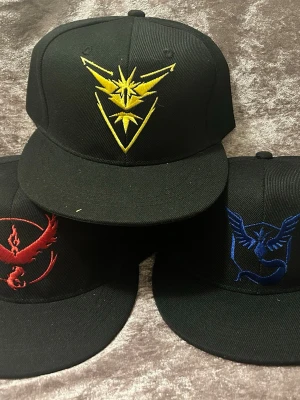 Svarta Pokémon Go kepsar med brodyr - Tre svarta snapback-kepsar med broderade Pokémon Go-lagloggor: gul för Team Instinct, röd för Team Valor och blå för Team Mystic. Kepsarna har platt skärm och justerbar baksida. Perfekt accessoar för Pokémon-fans som vill visa sitt lag. 145kr/st 