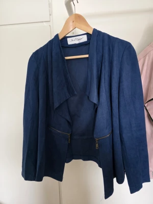 Blå draperad jacka från Jus d'Orange  - Snygg blå jacka från Jus d'Orange med draperad front och två dekorativa dragkedjor. Jackan har lång ärm och är tillverkad i ett mjukt, mockaliknande material. Perfekt för att lyfta vilken outfit som helst med en modern touch.