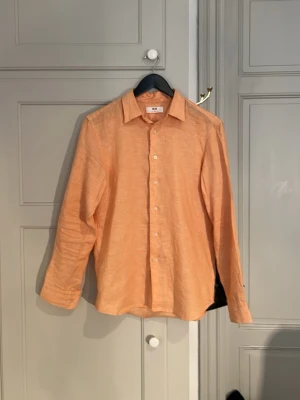Ljus orange linneskjorta från Uniqlo - Snygg ljus orange/ lax orange skjorta från Uniqlo i 100% linne. Klassisk krage, långärmad och med knappar framtill. Perfekt för dig som gillar en avslappnad men ändå stilren look. Skjortan har en lätt och luftig känsla som passar till många outfits. Perfekt till sommaren när det är varmt. Storlek XS, nypris 600kr