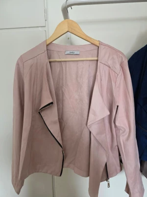 Ljusrosa skinnjacka från be-fun - Snygg ljusrosa skinnjacka från be-fun med asymmetrisk dragkedja och mjukt, följsamt material. Jackan har långa ärmar och en öppen, draperad front som ger en trendig look. Perfekt för att lyfta din outfit med en feminin touch.