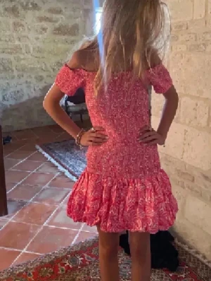 Rosa offshoulder kortklänning - Söt rosa kortklänning med vitt bladmönster och volang nertill. Klänningen har offshoulder-modell och är figurnära upptill med en luftig kjol. Perfekt för varma dagar och har en ungdomlig vibe.