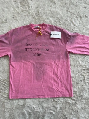 Rosa långärmad t-shirt Acne Studios - Rosa långärmad t-shirt från Acne Studios med tryck 'Acne Studios STOCKHOLM 1996' på bröstet. Klassisk rund halsringning och rak passform. Perfekt för dig som gillar stilrena och trendiga plagg med en touch av streetwear.