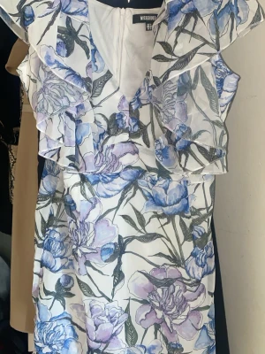 Blommig volangklänning från Missguided - Superfin vit klänning från Missguided med blått och lila blommönster, volangdetaljer vid axlarna och v-ringning. Klänningen har en feminin passform och är perfekt för vår och sommar. Materialet är lätt och känns bekvämt mot huden.