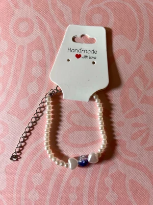 Pärlarmband med hjärtan och blå detalj - Sött handgjort armband med vita pärlor, två vita hjärtformade pärlor och en blå-glittrig pärla i mitten. Justerbar kedja i silverfärg. Perfekt accessoar för att lyfta din stil.