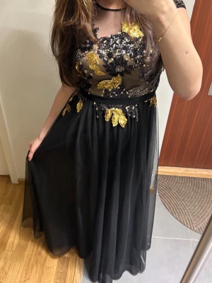 Bal/bröllops klänning - Svart långklänning med transparent överdel och detaljerade blommiga broderier i guld och svart. Klänningen har rund halsringning, korta ärmar och ett flytande kjolparti i tunt tyg. Perfekt för speciella tillfällen och passar perfekt till bal. Använd max 2 gånger. Är 170 cm lång