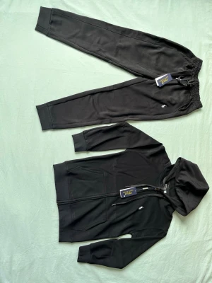 Svart loungewear-set från Polo Ralph Lauren - Svart loungewear-set från Polo Ralph Lauren med mjuk huvtröja och matchande byxor. Tröjan har dragkedja, huva med dragsko och två fickor framtill. Byxorna har resår i midjan och smala benslut. Båda plaggen har den klassiska Polo-loggan broderad.