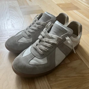 Maison margiela gats - Snygga sneakers i vitt och ljusgrått med ovandel i läder och mocka. Klassisk låg modell med bruna gummisulor och vita skosnören. Perfekta för en clean och stilren look. Utan box