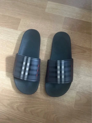 Svarta Burberry tofflor med rutmönster - Snygga svarta tofflor från Burberry med klassiskt rutmönster i grått, vitt och rött på remmen. Tillverkade i syntetmaterial med en bekväm, formad sula. Perfekta för en chill och trendig look.