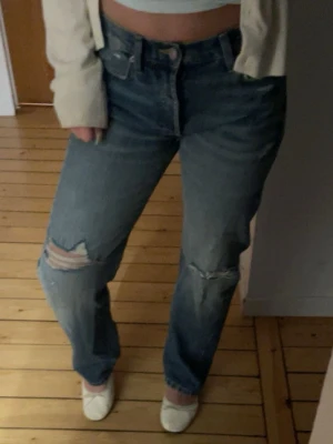 Ljusblå slitna raka jeans - Säljer ett par ljusblå jeans med raka ben och slitningar vid knäna och låret. Klassisk femficksmodell med hög midja och knappgylf. Jeansen har en avslappnad passform och är tillverkade i denim av bomull. Knappt använda, säljer för att de är för små