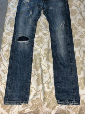 Blå slitna jeans med patch och detaljer - Säljer ett par blå jeans med slitningar, hål och patch på knät. Jeansen har raka ben och klassiska femficksdesign. På baksidan finns en dekorativ patch på ena fickan. Materialet är denim och färgen är tvättad blå.