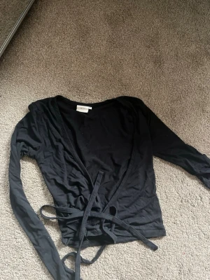 Svart omlottopp från Nelly XS - Svart långärmad omlottopp från Nelly med knytband i midjan. Tunn och stretchig i materialet, perfekt för lager-på-lager. Enkel och stilren design som är lätt att matcha med olika outfits.