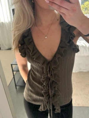 Ribbad volangtopp från Zara - Supersöt ribbad topp från Zara i khaki med volangdetaljer längs v-ringningen och ärmarna. Toppen har knyt framtill och är ärmlös, perfekt för att lyfta vilken outfit som helst.