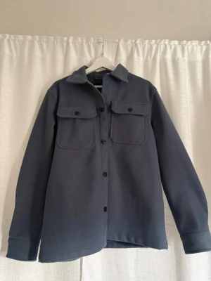 Mörkblå overshirt med fickor - Stilren mörkblå overshirt med klassisk krage och två stora bröstfickor med lock och knappar. Rak passform och knäppning framtill. Perfekt lager-på-lager-plagg för en avslappnad look.