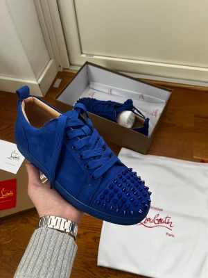 Blåa sneakers från Christian Louboutin - Snygga blåa sneakers från Christian Louboutin i mocka med matchande blå snörning och coola nitar på tån. Skorna har en låg siluett och exklusiv känsla med beige innersula och klassisk Louboutin-logga. Perfekta för dig som vill sticka ut med din stil.