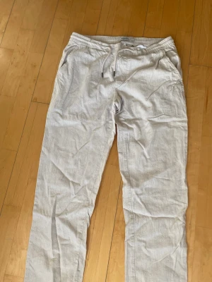 Ljusgrå linnebyxor med dragsko - Snygga ljusgrå linnebyxor med elastisk midja och dragsko. Byxorna har sidfickor fram och två bakfickor med knapp. Perfekta för en avslappnad och stilren look. Tillverkade i svalt linnematerial.
