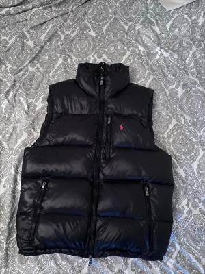 Svart dunväst från Polo Ralph Lauren - Svart dunväst från Polo Ralph Lauren med hög krage och två dragkedjeförsedda fickor. Västen har ett quiltat mönster och en liten röd logotyp på bröstet. Perfekt för att hålla värmen under kyliga dagar.