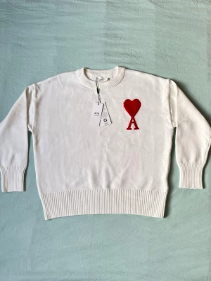 Vit stickad tröja från Ami Paris - Vit stickad tröja från Ami Paris med rund halsringning och ribbade muddar. På bröstet finns ett rött hjärta med bokstaven A, vilket är varumärkets ikoniska logga. Tröjan är i mjukt material och har en avslappnad passform.